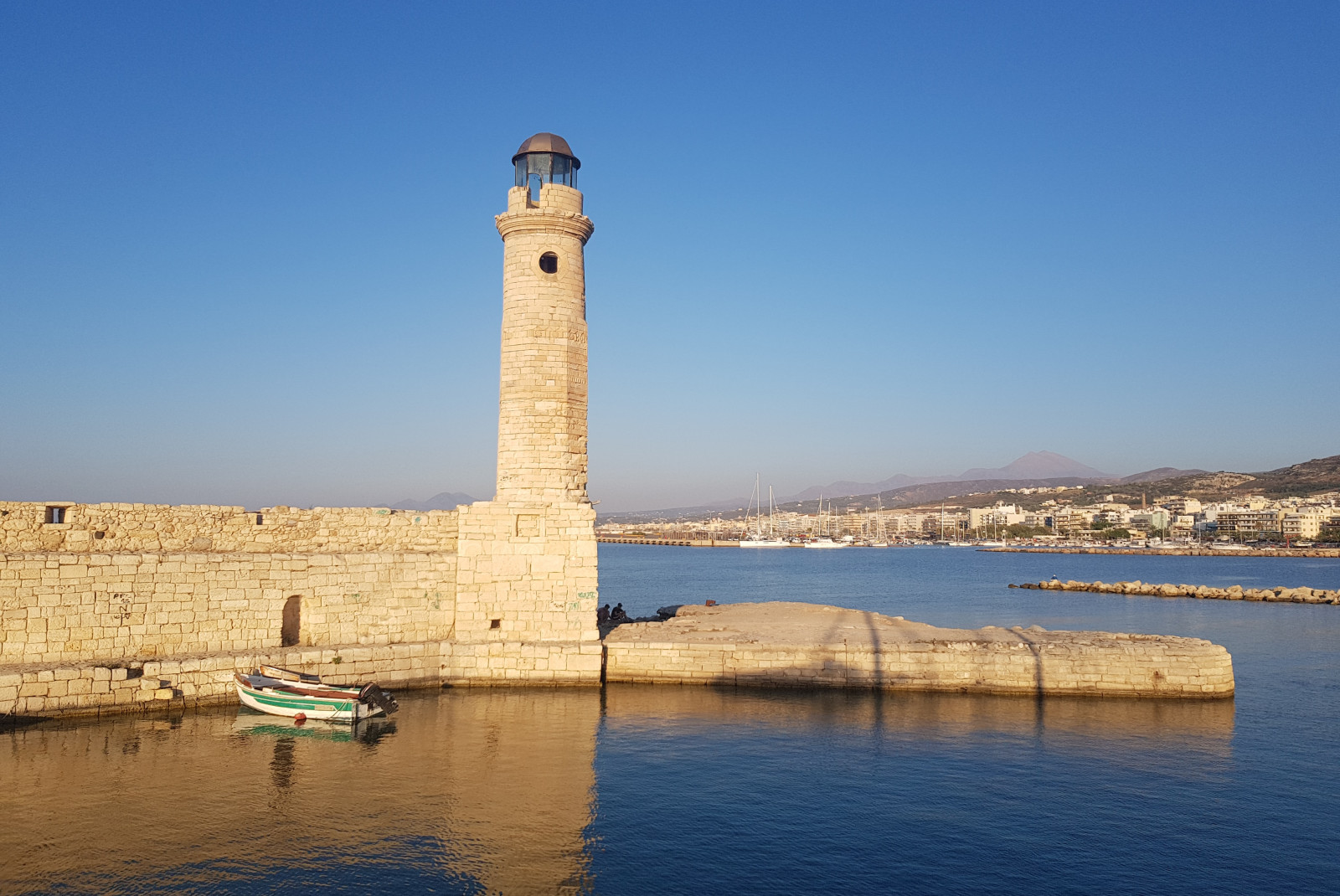 Stone white lighthouse with blue water and sky and a small blue boat and seaside towns and mountains in the background