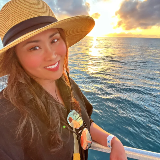 Travel Advisor Cheska Tariga leans on the railing of a boat cruising through the sunset wearing a sun hat