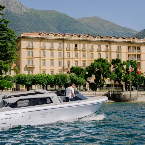 A white boat on Lake Como in front of a luxurious yellow hotel with green trees and mountains.