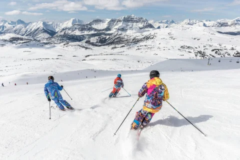 Three people skiing downhill in colorful outfits with snowy mountains in the background.