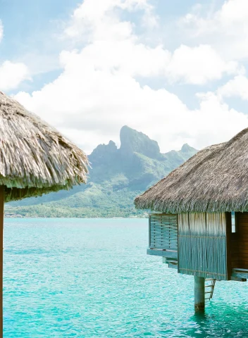 Overwater bungalows with the mountains, sky, and clear, blue water.
