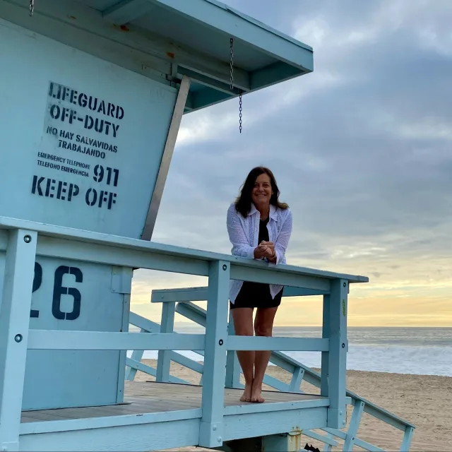 Picture of Deb wearing a white top and standing on a life guard tower turned against the beach and a sunset over the water