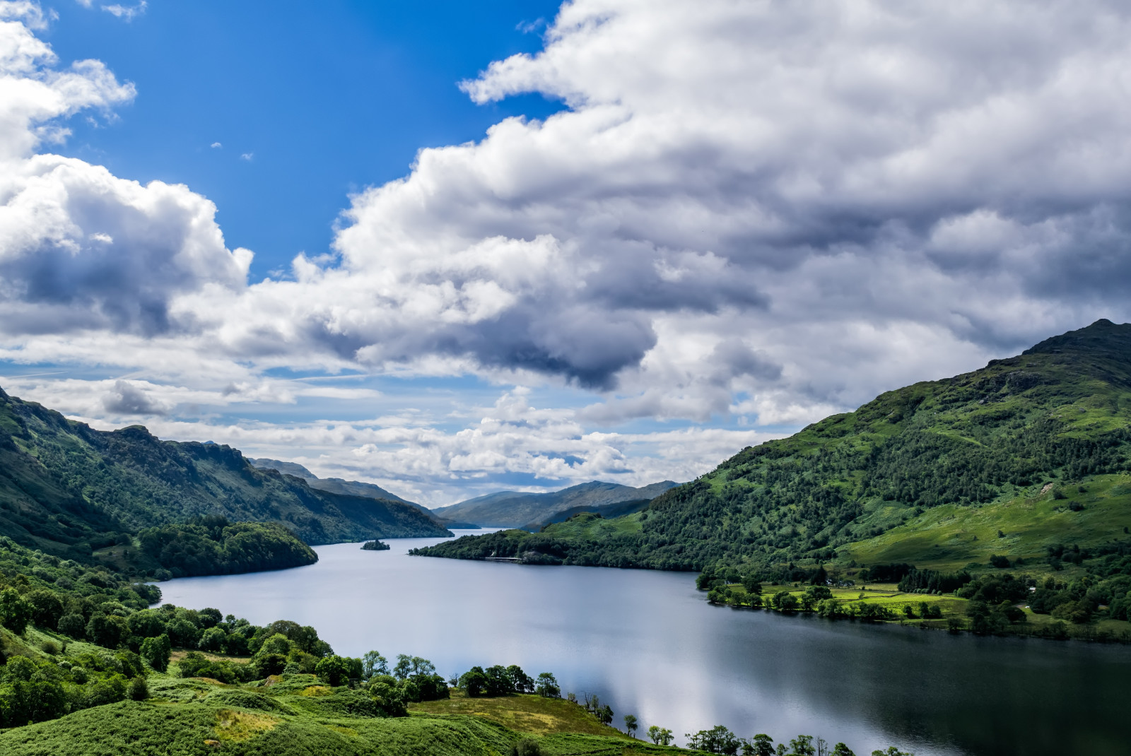 Blue lake surrounded by green mountains with clouds and sun in the sky