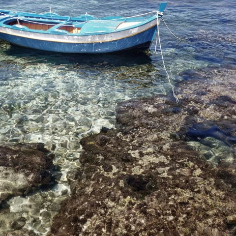 A small fishing boat drifting in the beautiful crystal clear water off the shore with rocks. 