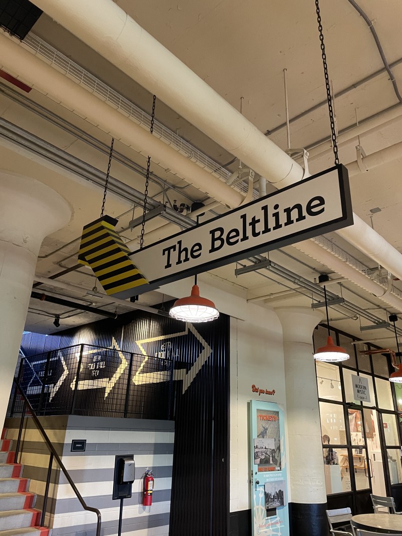 A black, yellow and white sign that reads 'The Beltline' in a building with striped walls and white industrial ceilings.