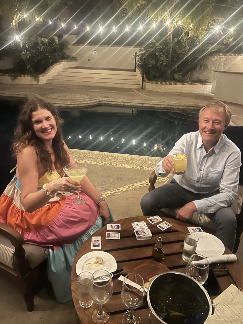 A woman and a man sitting at a table enjoying a drink and playing a card game by a pool area with string lights in the evening