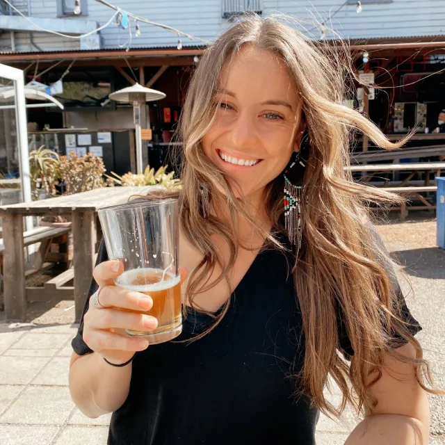 Amanda posing with a drink in hand while wearing a black t-shirt in front of a restaurant patio