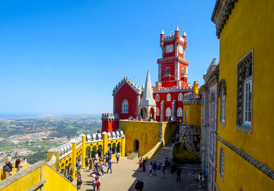 people walk around a yellow and orange castle atop a city on a clear blue day
