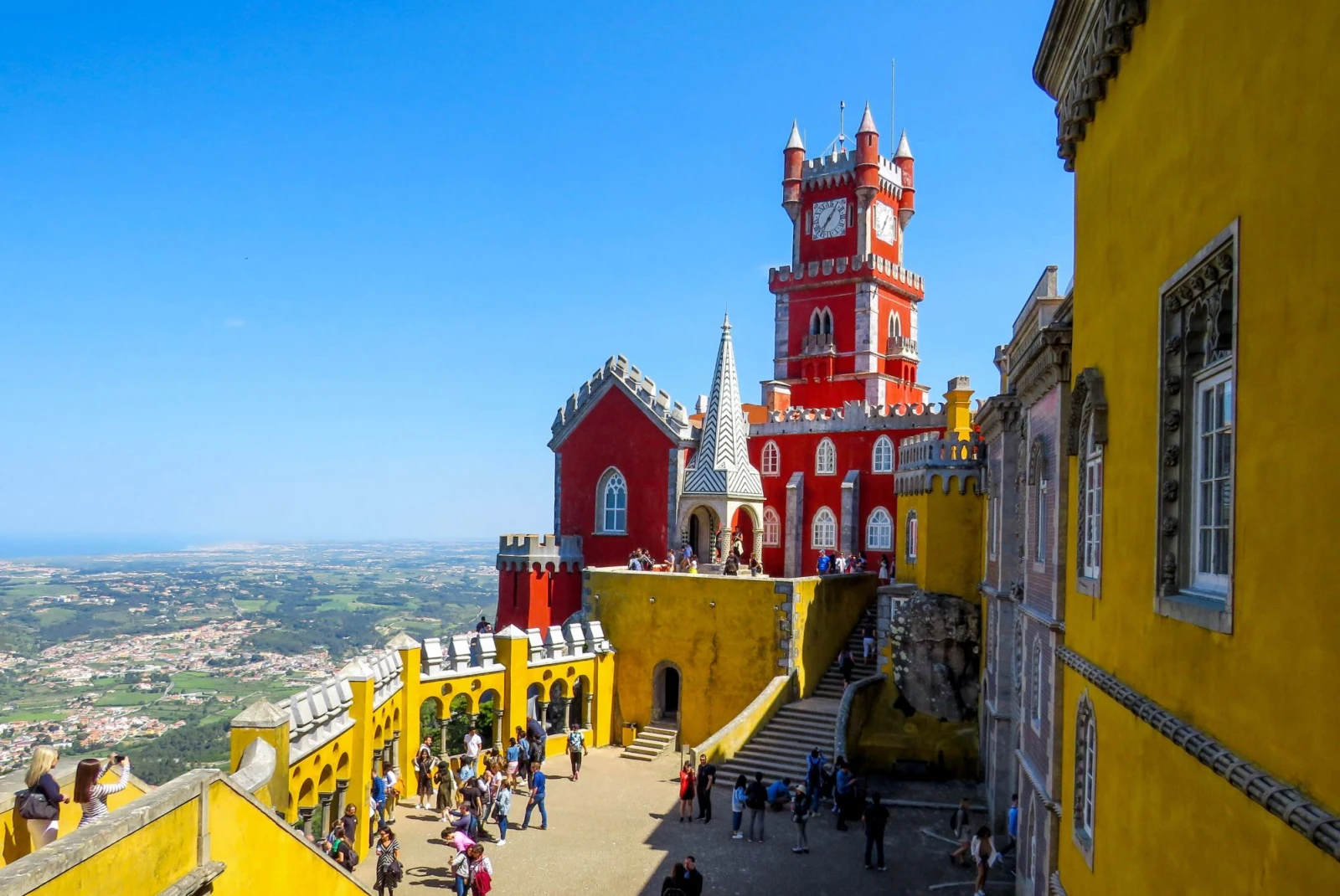 people walk around a yellow and orange castle atop a city on a clear blue day