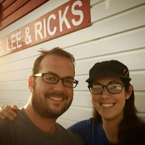 A couple posing for a photo in front of a white building with a red sign that reads Lee & Ricks. 
