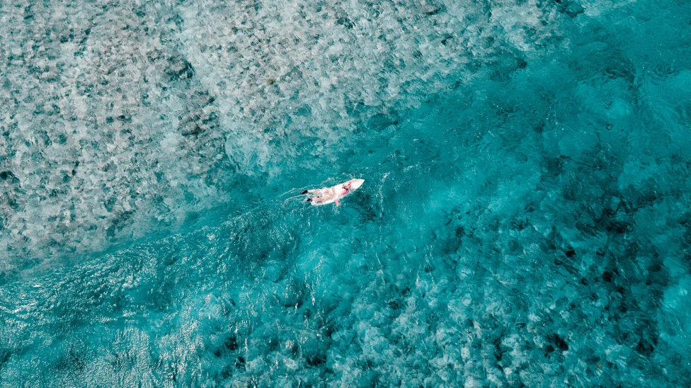 A woman in white on a white surfboard in crystal blue waters with white sand for a luxury surf vacation in the Maldives.