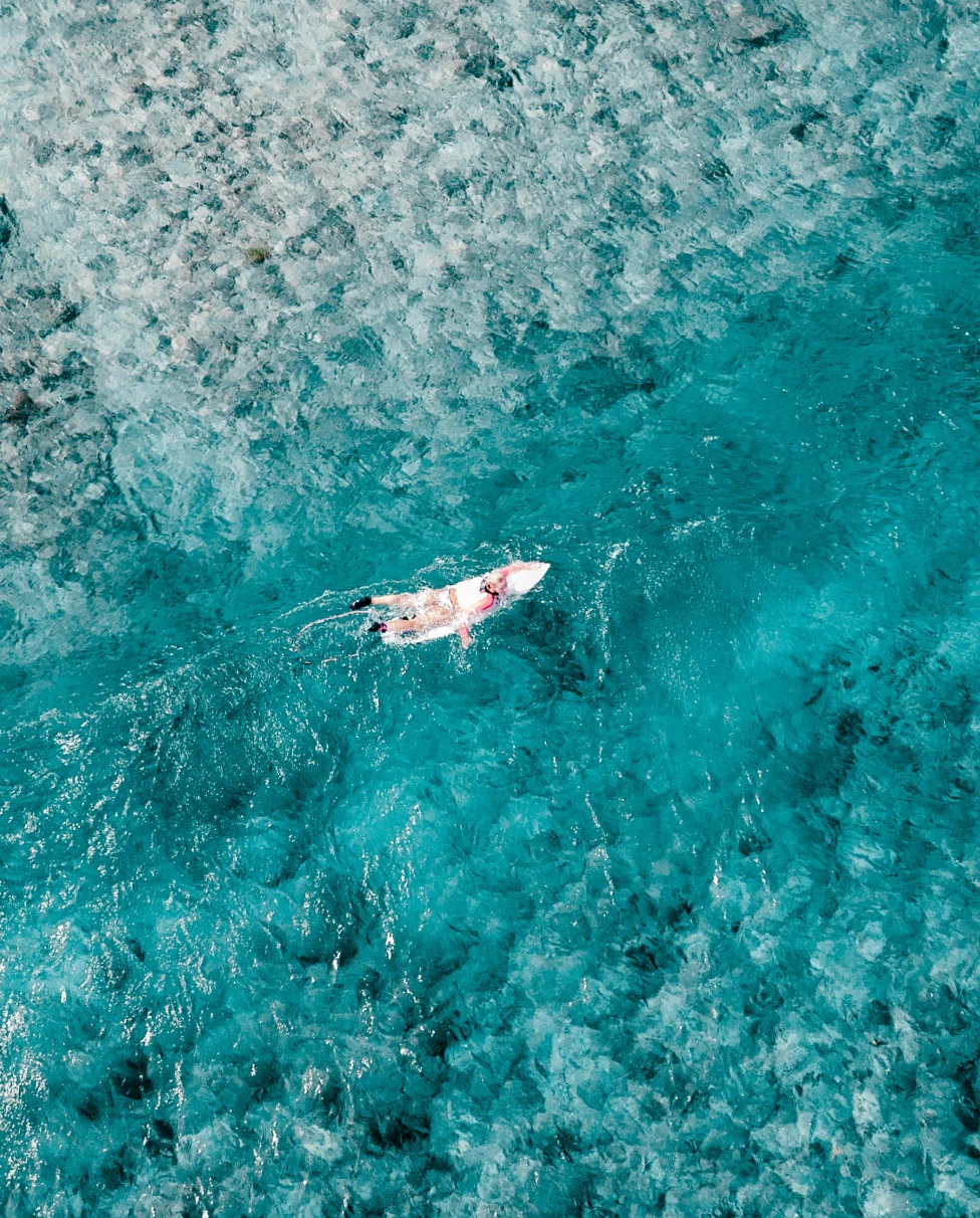 A woman in white on a white surfboard in crystal blue waters with white sand for a luxury surf vacation in the Maldives.