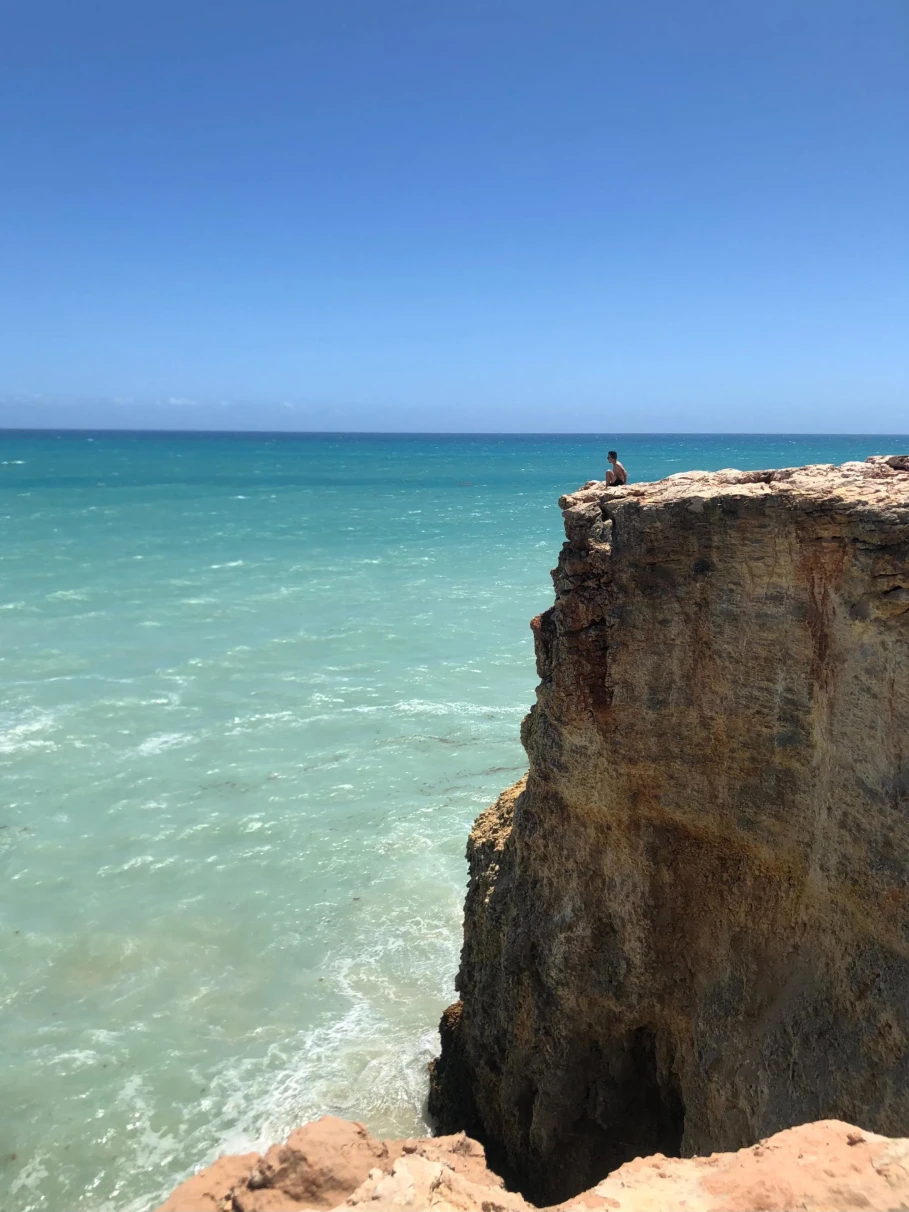 a person sits on the edge of a high stone cliff looking out at the clear light blue ocean on a cloudless day