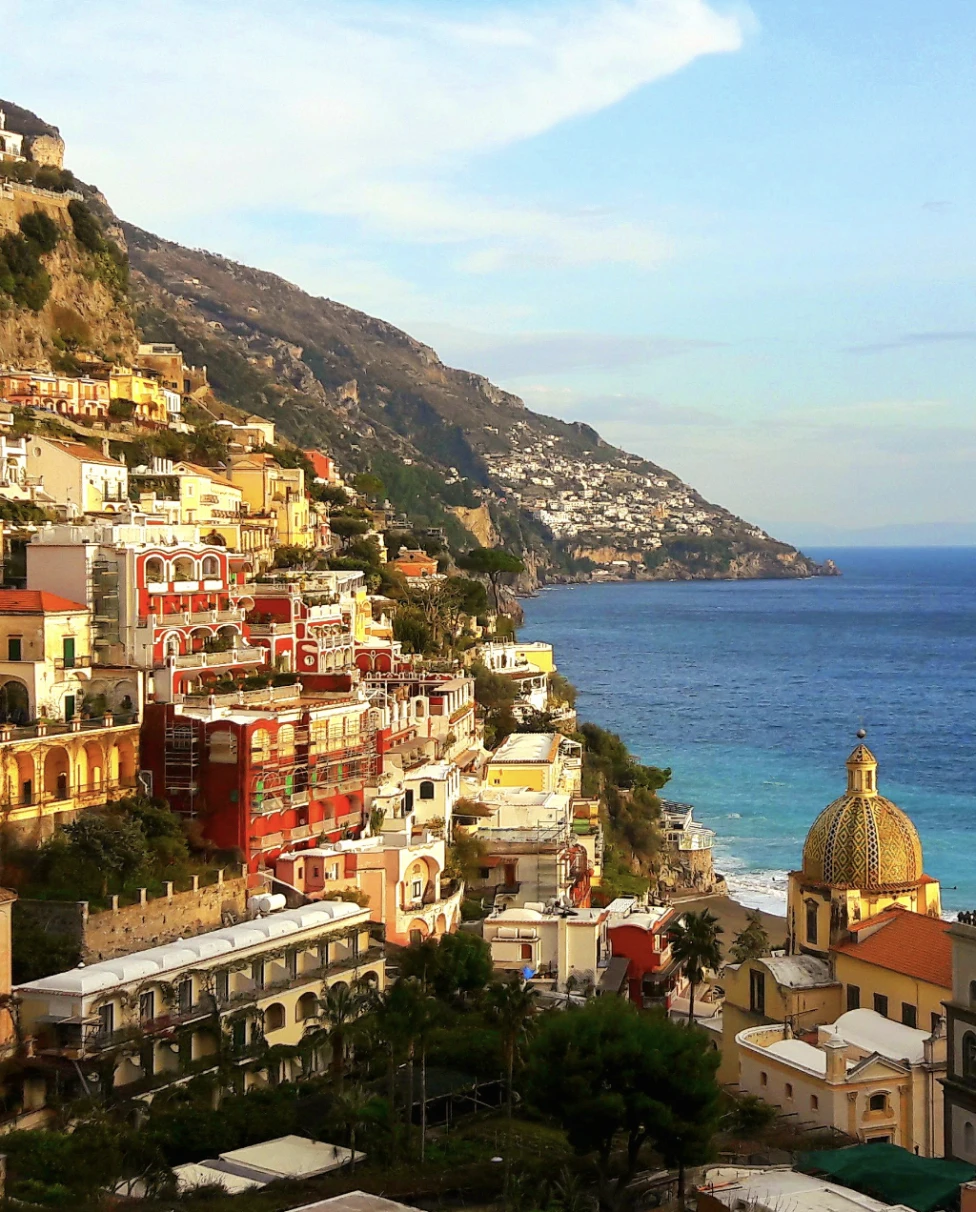 A picture of the vibrantly colorful town of Positano, with various buildings nestled into a mountain. There is also a view of the blue sea in the background towards the right of the image. 