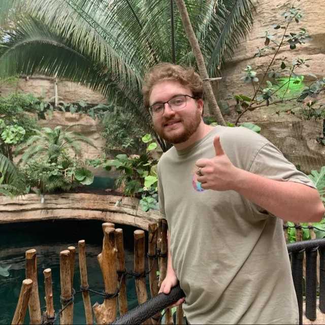 Ryan wearing a light green tee shirt and holding a thumbs up hand signal while standing in front of a rock wall and palm tree