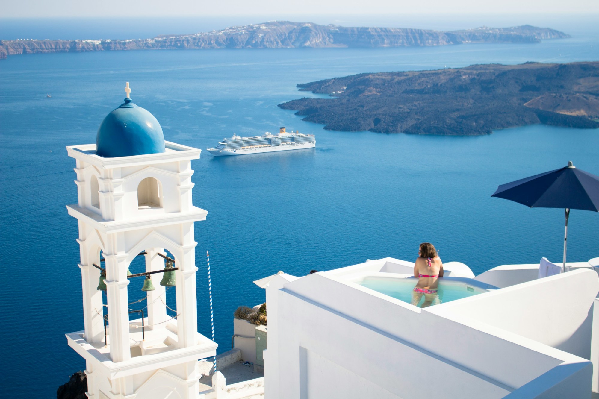 A woman looks out from a pool at a whitewashed Greek seaside resort toward the sea as a cruise ship passes by