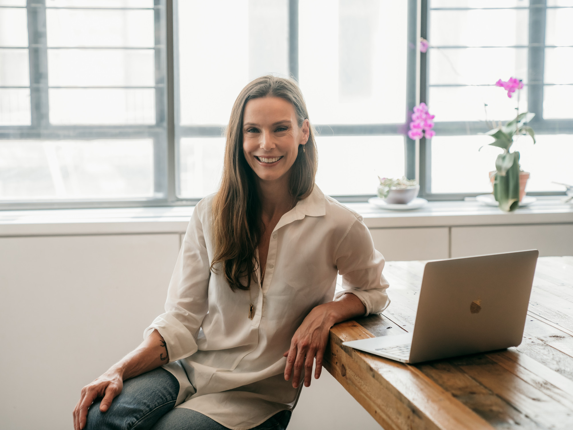A woman in a white shirt sits at a wooden table with her laptop