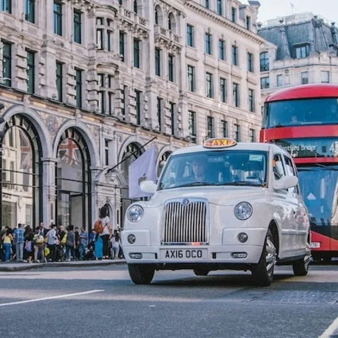 A red colored double decker and a white car on the road in London.