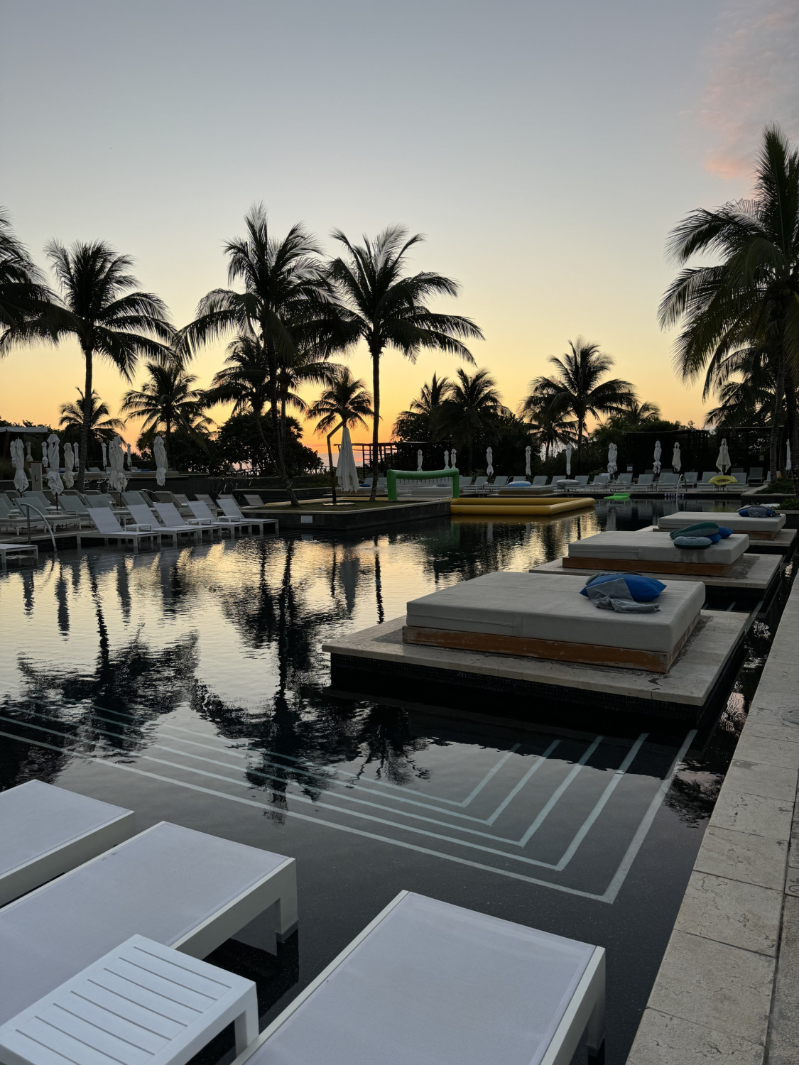 A swimming pool surrounded by lawn chairs, palm trees and day beds. There is a lush palm tree in the background and a golden sunset in the background. 