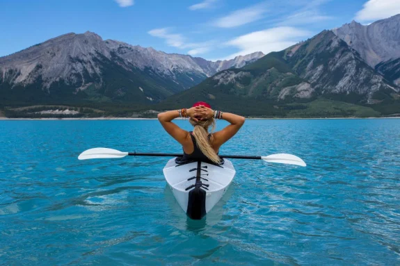 Girl wearing red hat sits in white kayak in bright blue water with the mountains in the background