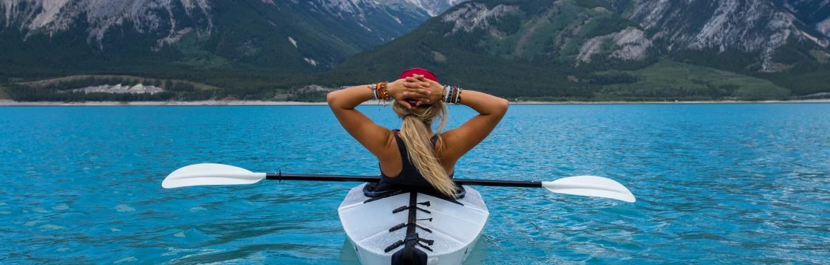 Girl wearing red hat sits in white kayak in bright blue water with the mountains in the background