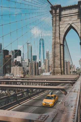 yellow taxi driving over large bridge with city skyline in the background