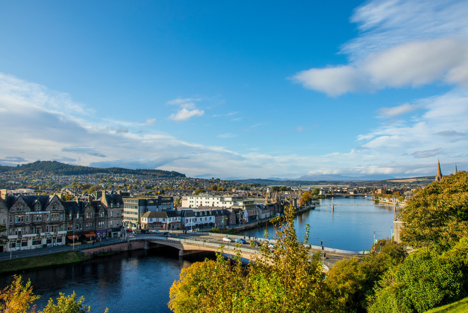 Overlooking the town of Inverness, Scotland with a cluster of brown and white tall homes and buildings, a bridge going over a blue river and trees at the basin.