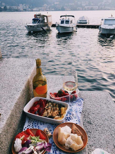 Dishes of bread and vegetables with a wine glass and bottle overlooking the water in Italy