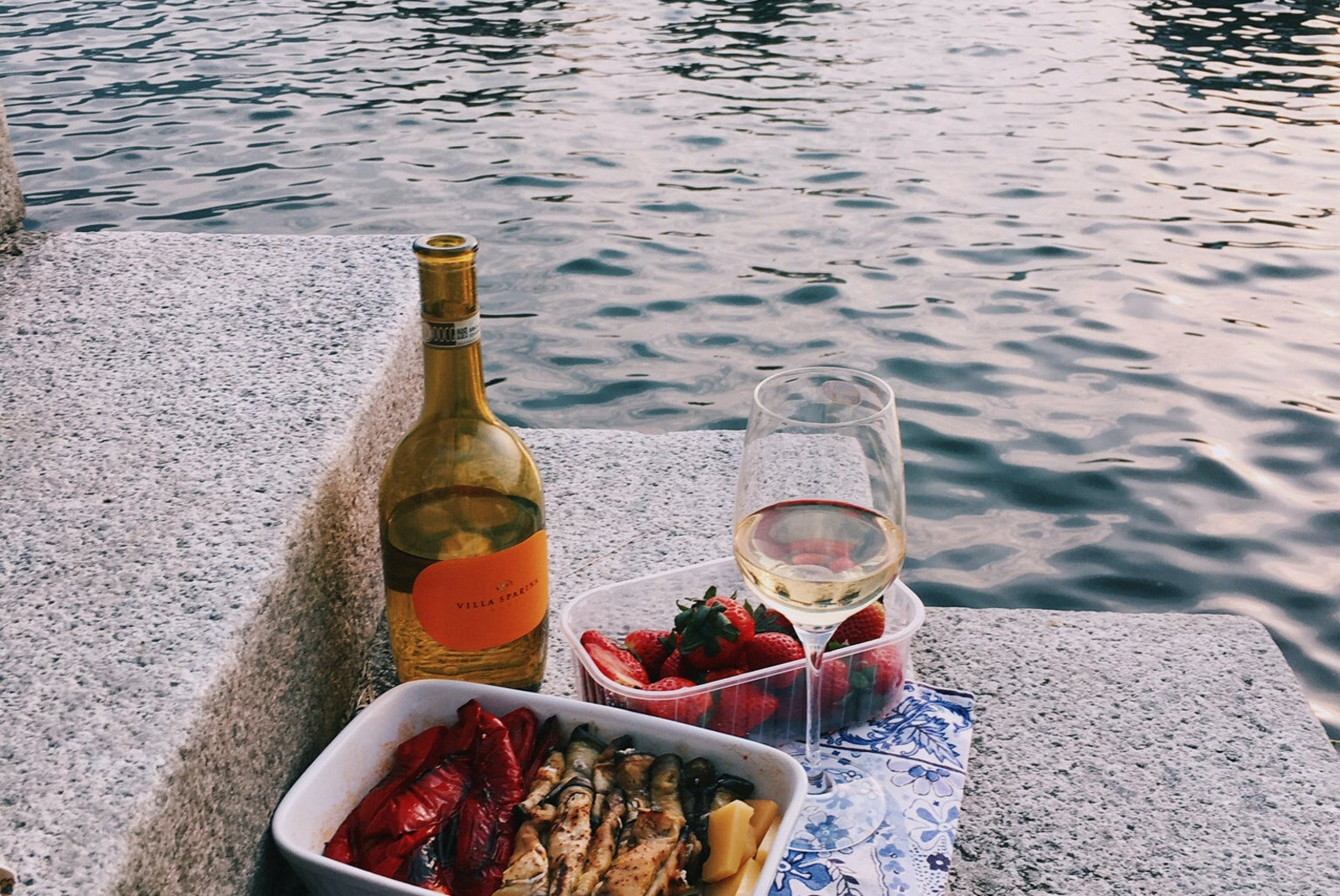 Dishes of bread and vegetables with a wine glass and bottle overlooking the water in Italy