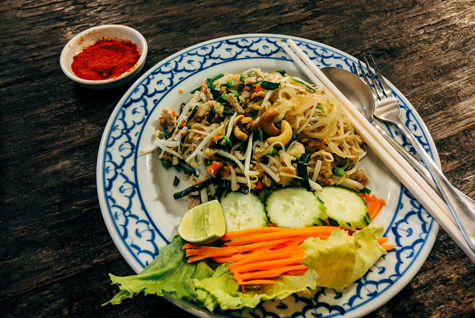 Pad Thai on a blue and white plate with red ground pepper, tan chopsticks, silver spoon and fork, and green cucumbers and lettuce and orange carrots in Bangkok, Thailand.