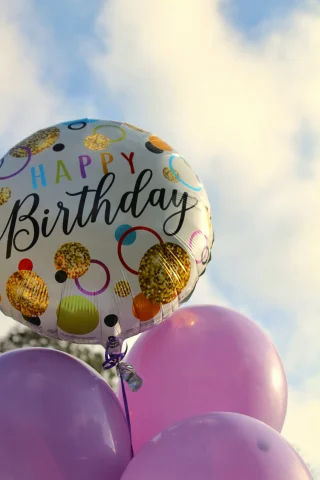 A group of four balloons, three of them being purple and the fourth one multicolored with text that reads "Happy Birthday". There is a cloudy sky in the background. 