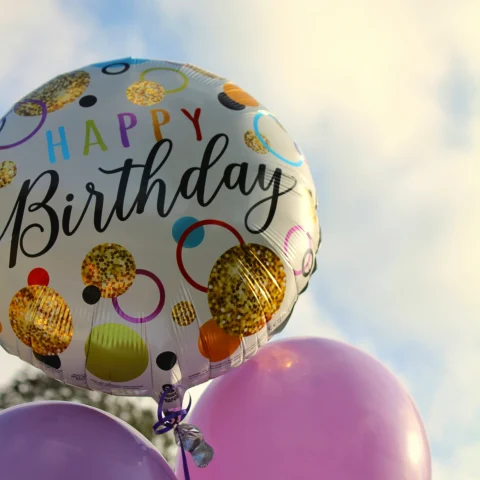 A group of four balloons, three of them being purple and the fourth one multicolored with text that reads "Happy Birthday". There is a cloudy sky in the background. 