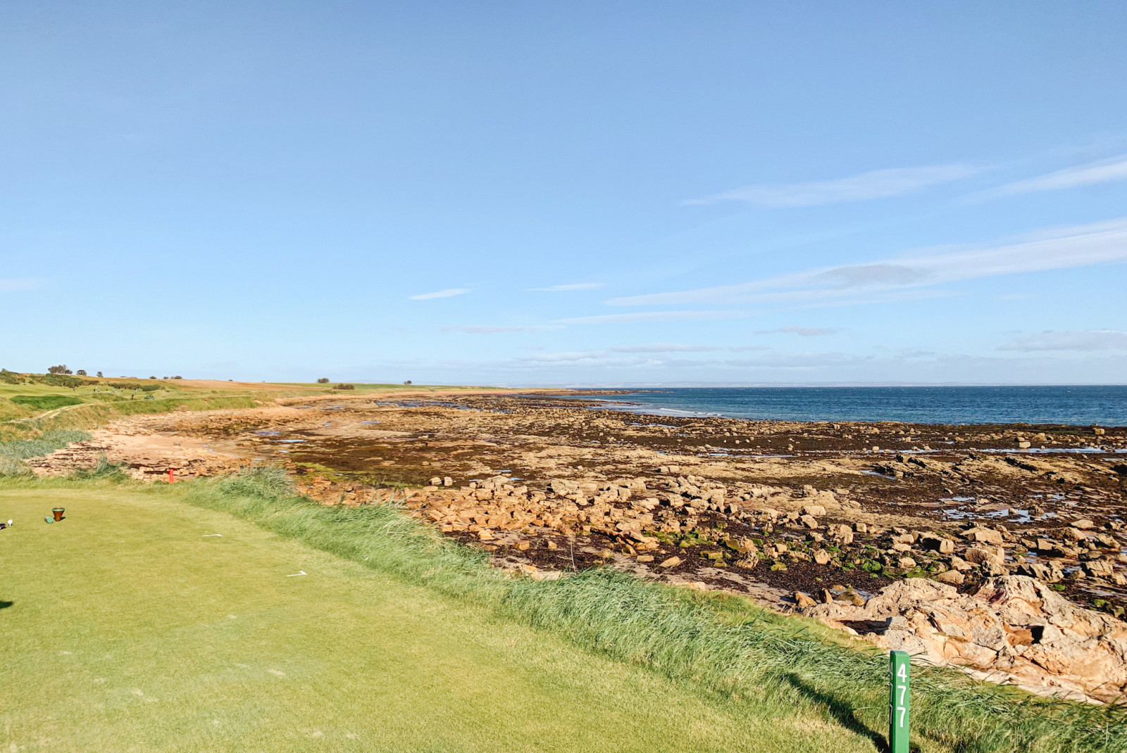 green grass golf course with a post that says 477 and tan rocks leading into blue ocean water
