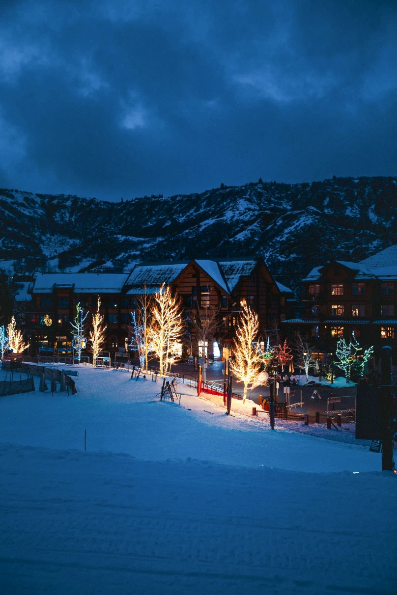 An early-evening scene of cabins, snow, mountains, and trees decorated with Christmas lights in Snowmass Village, Colorado.