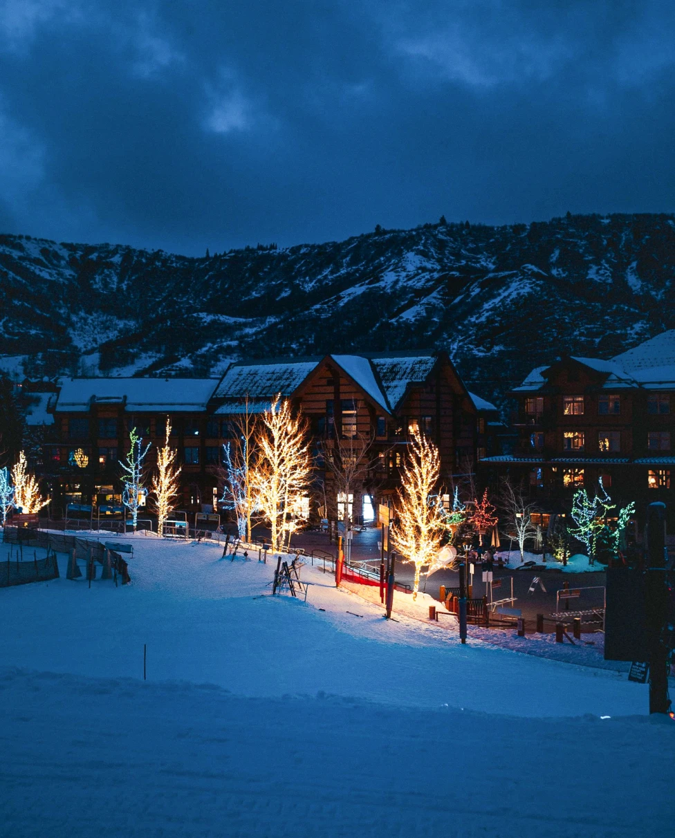 An early-evening scene of cabins, snow, mountains, and trees decorated with Christmas lights in Snowmass Village, Colorado.