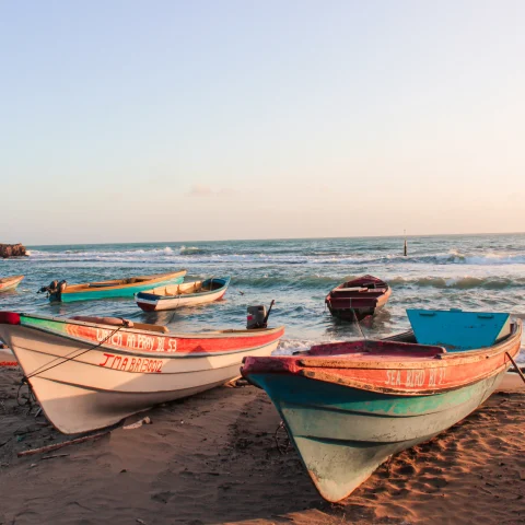 two white red and blue boats on a tan sandly beach at sunset with pink skies and blue ocean water with white crashing waves