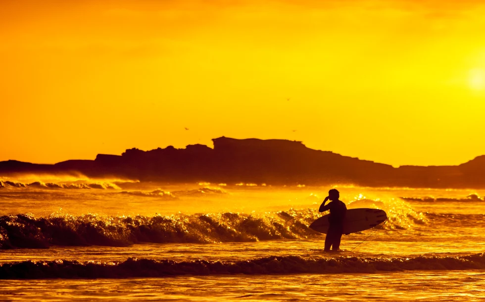 A black silhouette of a person carrying a surfboard with waves crashing and an orange sunset in Mexico.