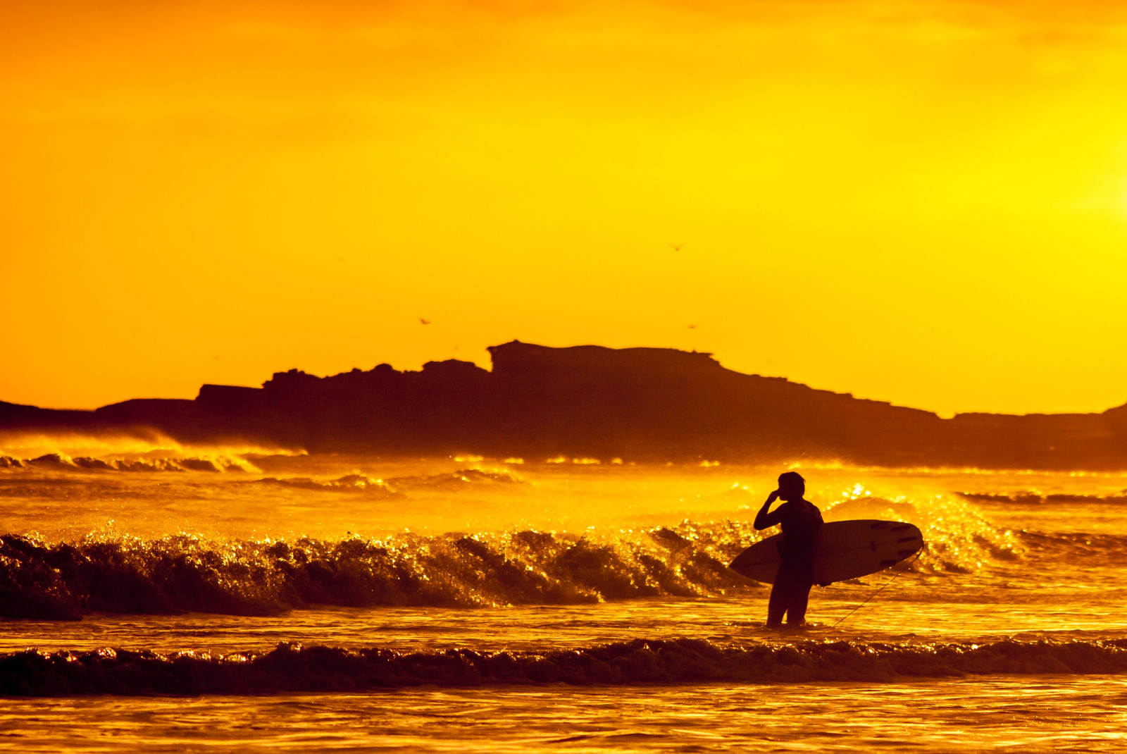 A black silhouette of a person carrying a surfboard with waves crashing and an orange sunset in Mexico.