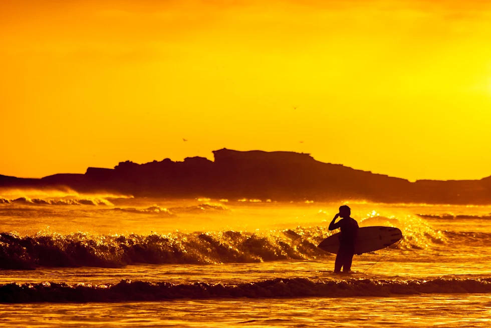A black silhouette of a person carrying a surfboard with waves crashing and an orange sunset in Mexico.