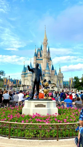 The Walt Disney and Mickey Mouse statue in front of the castle at Disney World. There are red flowers surrounding the statue and tourists walking throughout the surrounding areas. 