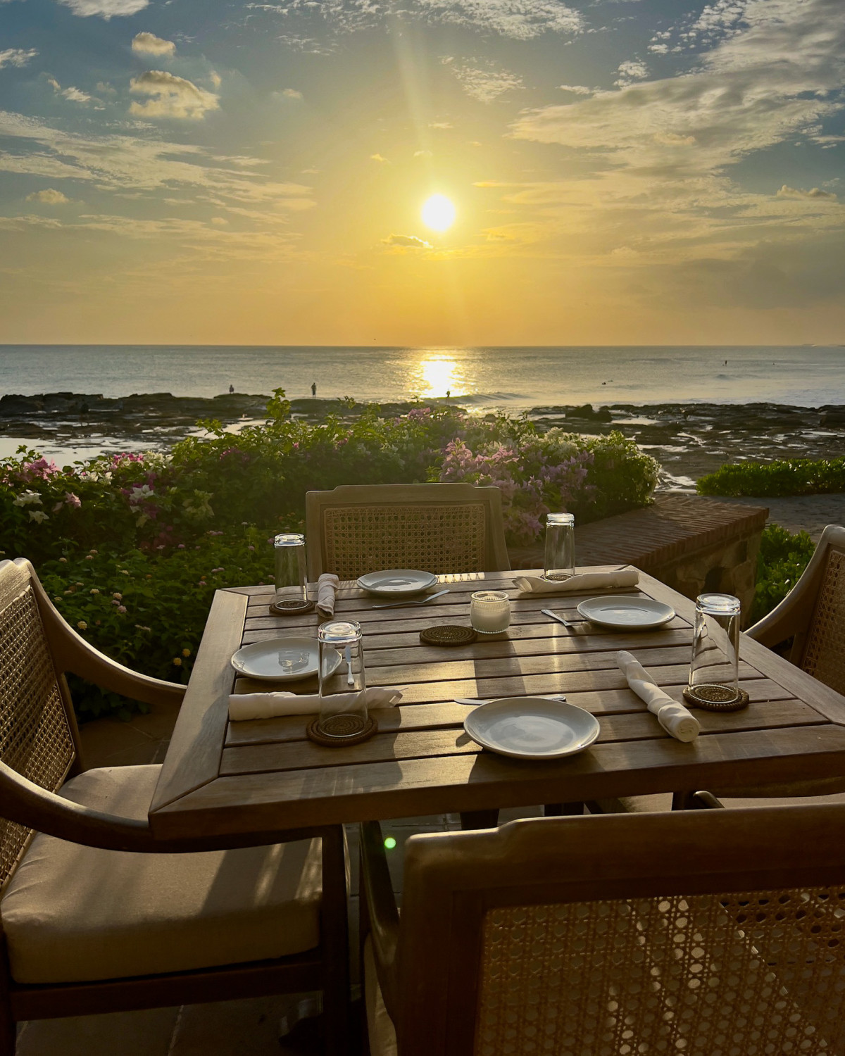 An outdoor dining area with a wooden table, set with plates, glasses and cutlery, overlooking the landscape and sea at sunset.