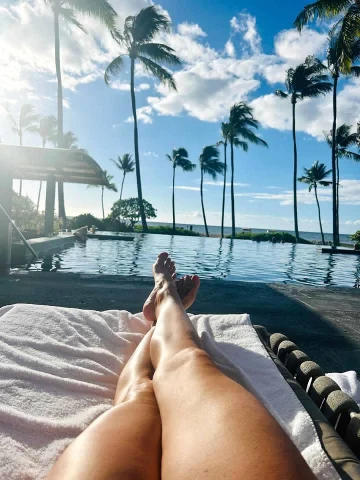 A pool with palm trees and legs of a person taking the picture lounging on pool chair.