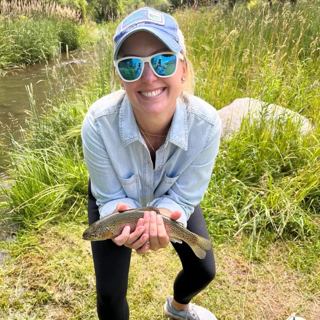 Genny holding a fish next to a pond surrounded by grass. 