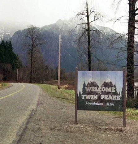 A roadside view of the "Welcome to Twin Peaks" sign, on a misty day, with a mountain peak in the background.
