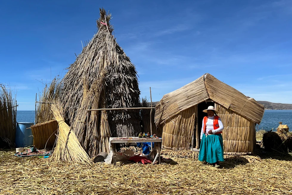 A lady in traditional dress standing in front of a lake side straw hut.