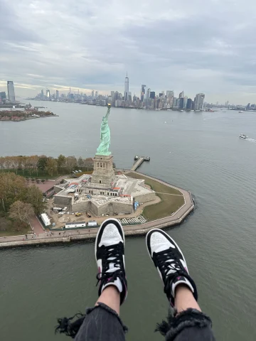 Travel Advisor's feet hanging off of an elevated ledge over the Hudson River, with Lady Liberty and New York City's skyline in the background.