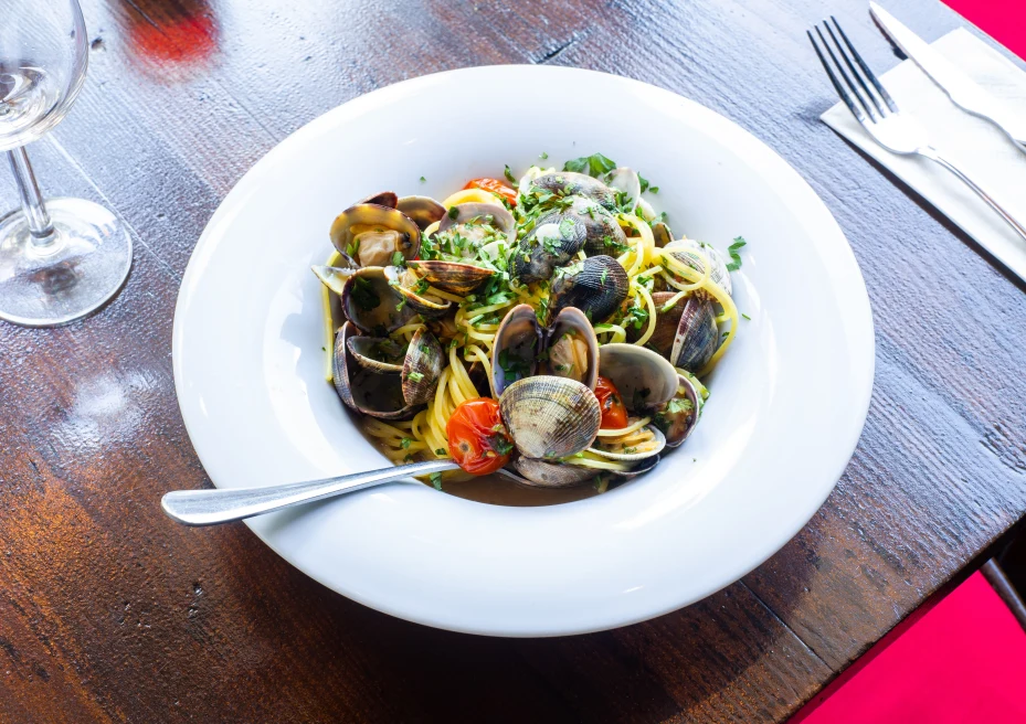White bowl of pasta and clams on a wooden table with silverware and wine glass