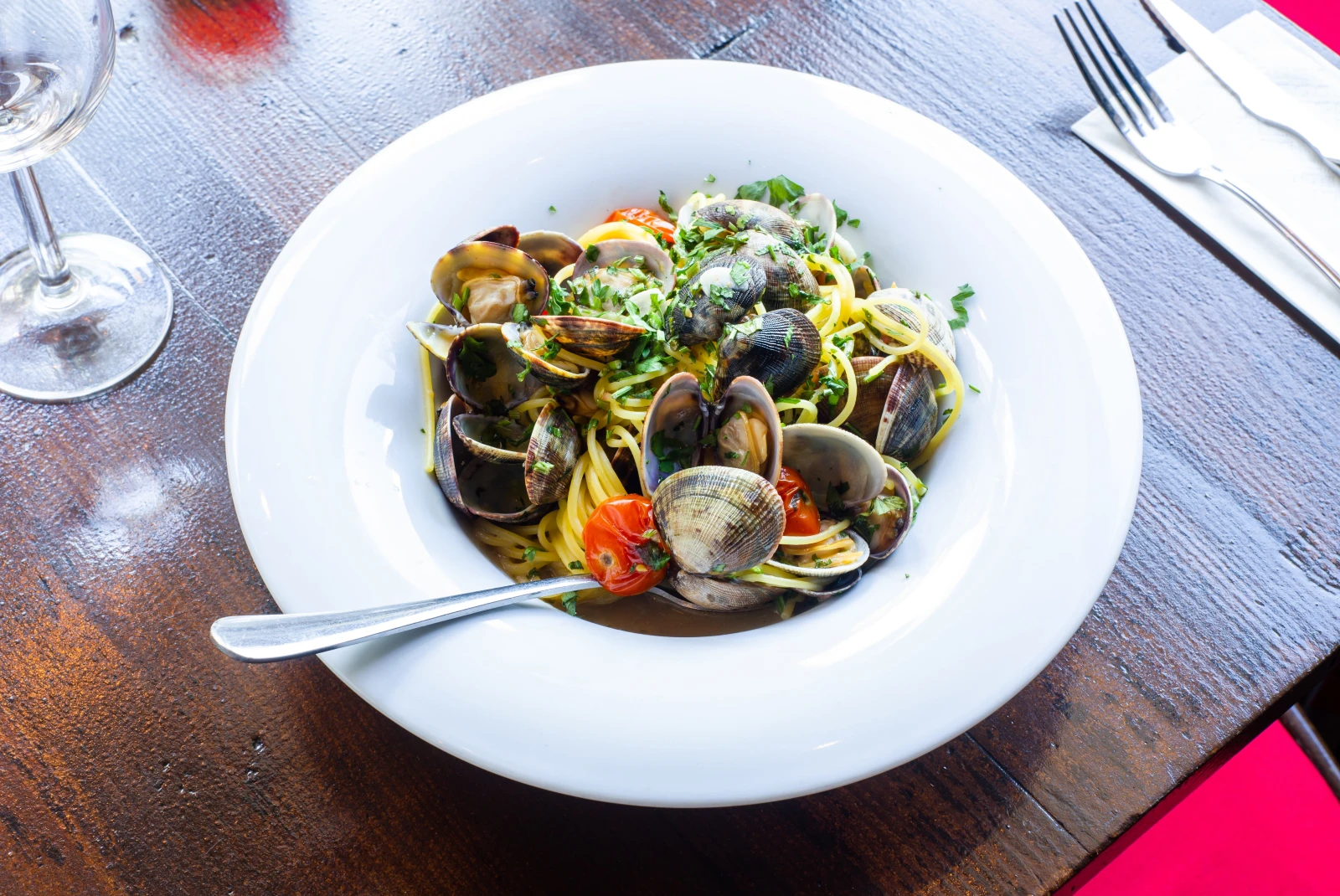 White bowl of pasta and clams on a wooden table with silverware and wine glass