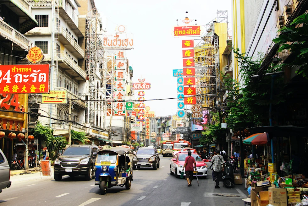 A crowded street full of cars, people, and tuk tuks in Bangkok, Thailand with yellow and red signs, white buildings, with green, leafy trees and phone lines.