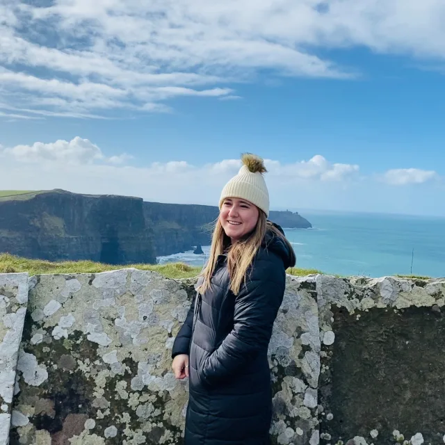 Travel Advisor Allison Albanese in a woolie hat and a long puffer jacket in front of sea. 